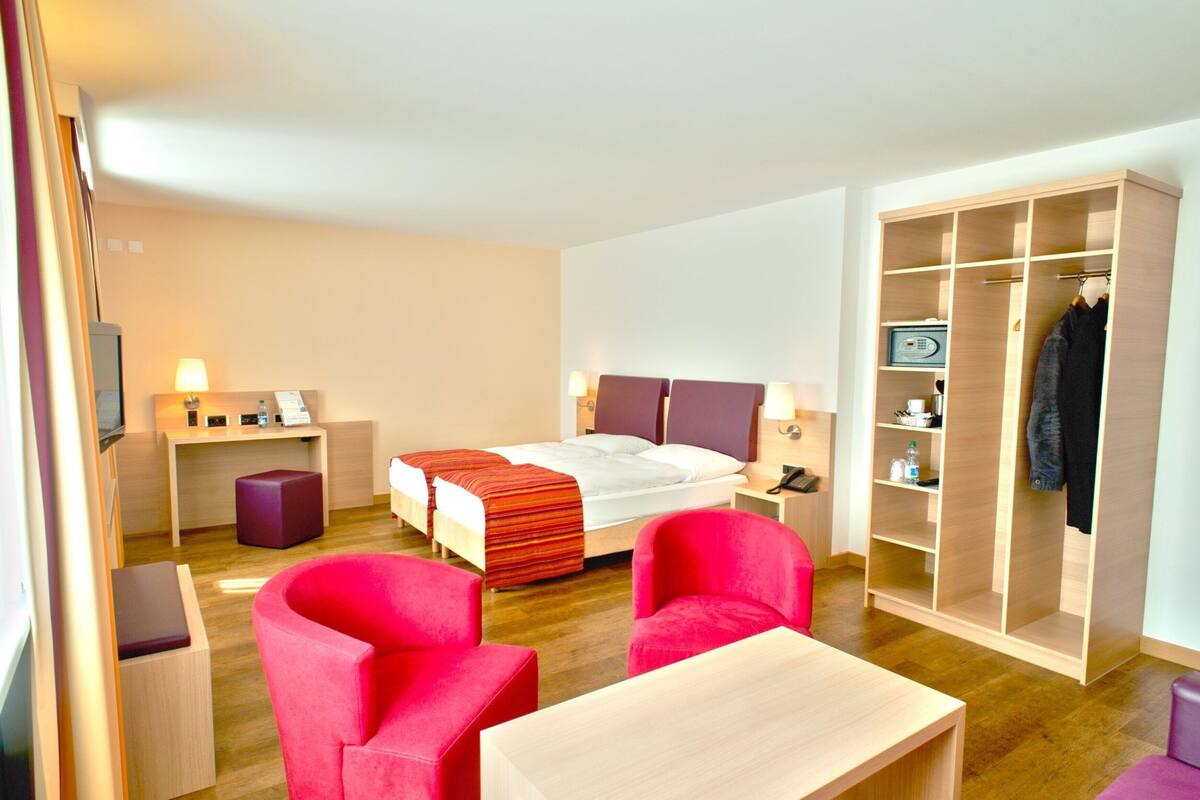 Ein Hotelzimmer mit Bett, zwei Nachttischen, einem Schreibtisch, einem Sessel, einem Kleiderschrank und einem Fernseher.