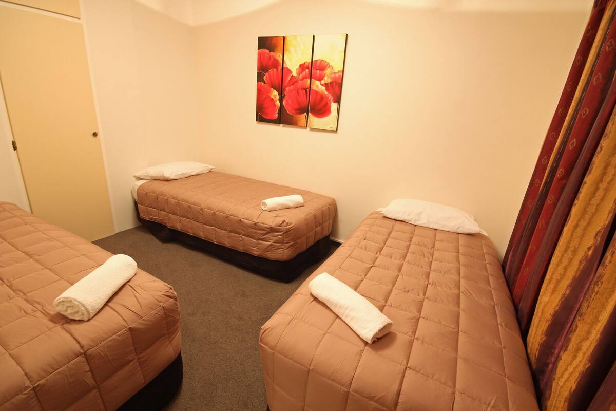 A hotel room with three beds, each with a brown quilted mattress and white pillows. There is a painting of red flowers on the wall.