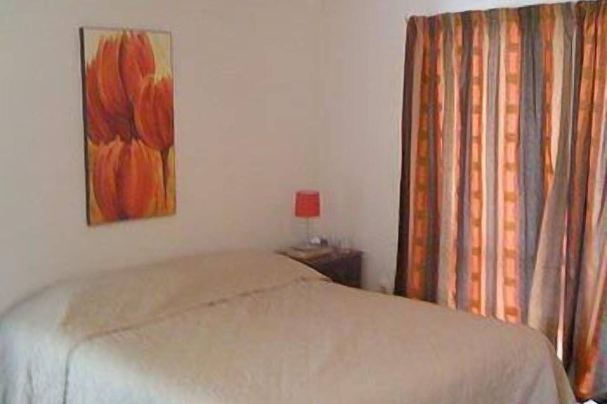 A hotel room with a bed, a painting on the wall, a nightstand with a lamp, and a window with curtains.