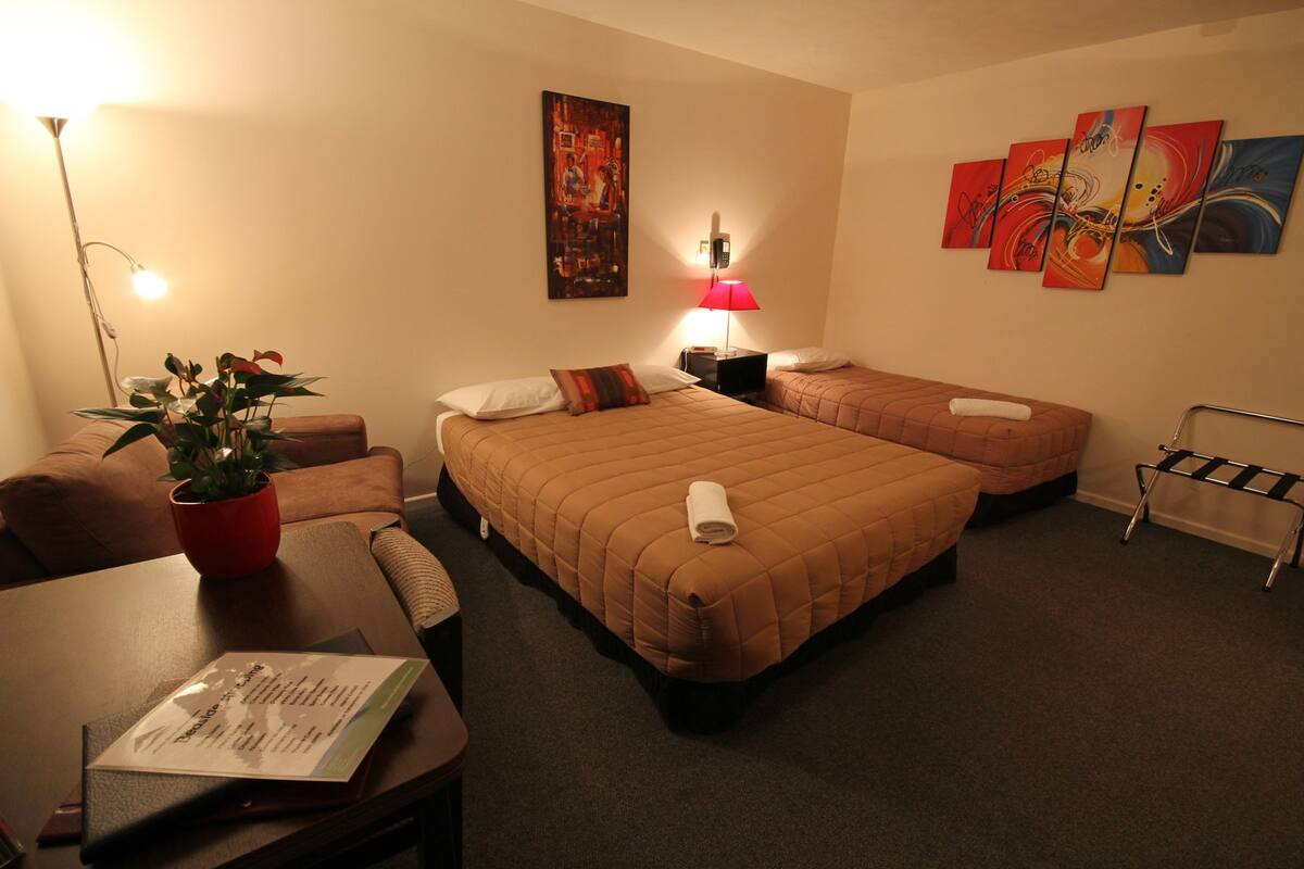 A hotel room with two beds, a desk, a chair, a plant, and artwork on the walls.