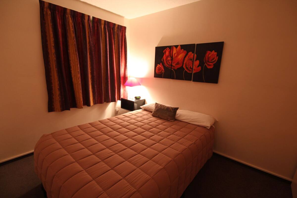 A hotel room with a large bed, a nightstand with a lamp, and a painting of red flowers on the wall.