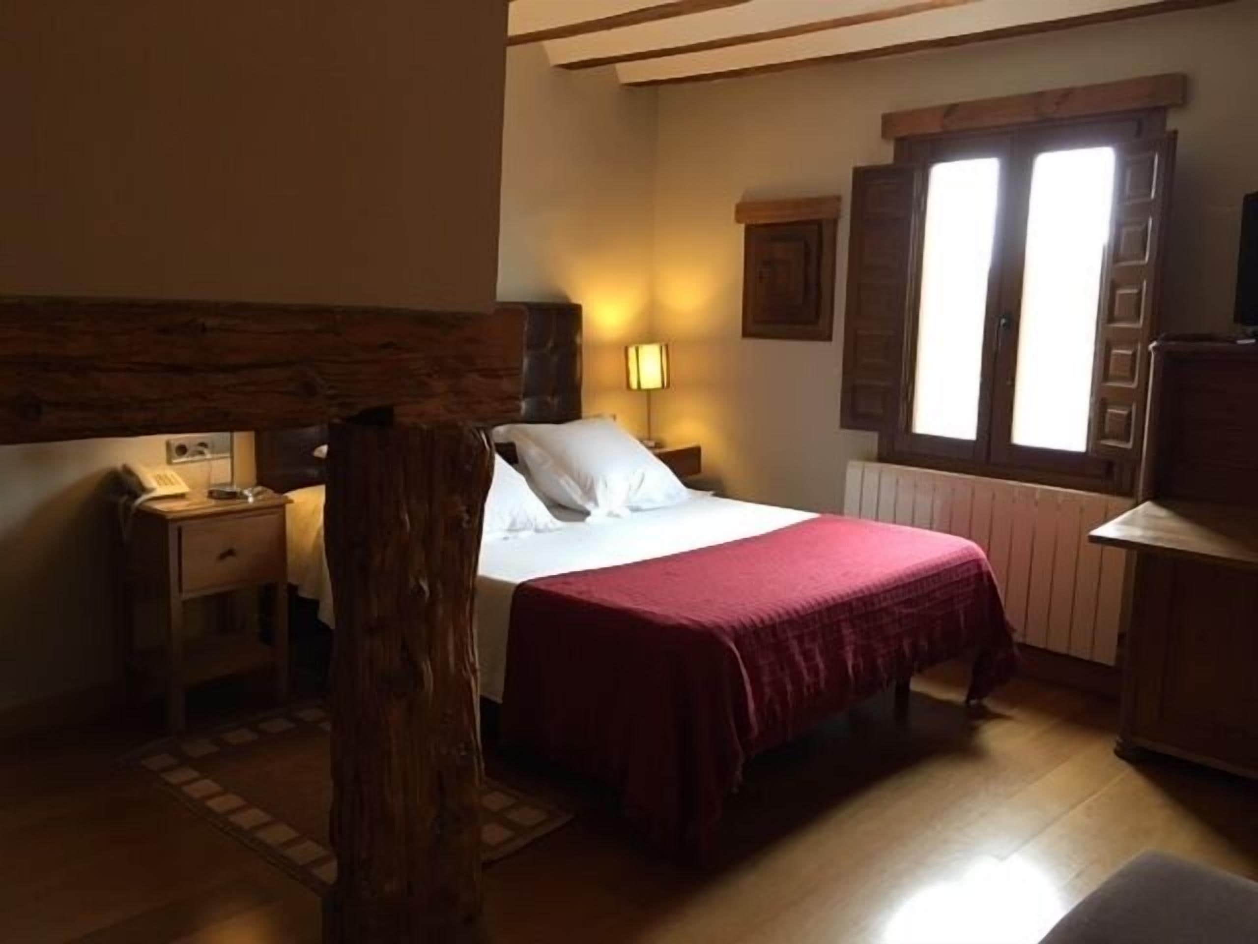 Ein Schlafzimmer mit Holz-Kopfteil, einem Bett mit roter Bettdecke, einem Nachttisch mit Lampe und einem Fenster mit Holzjalousien.