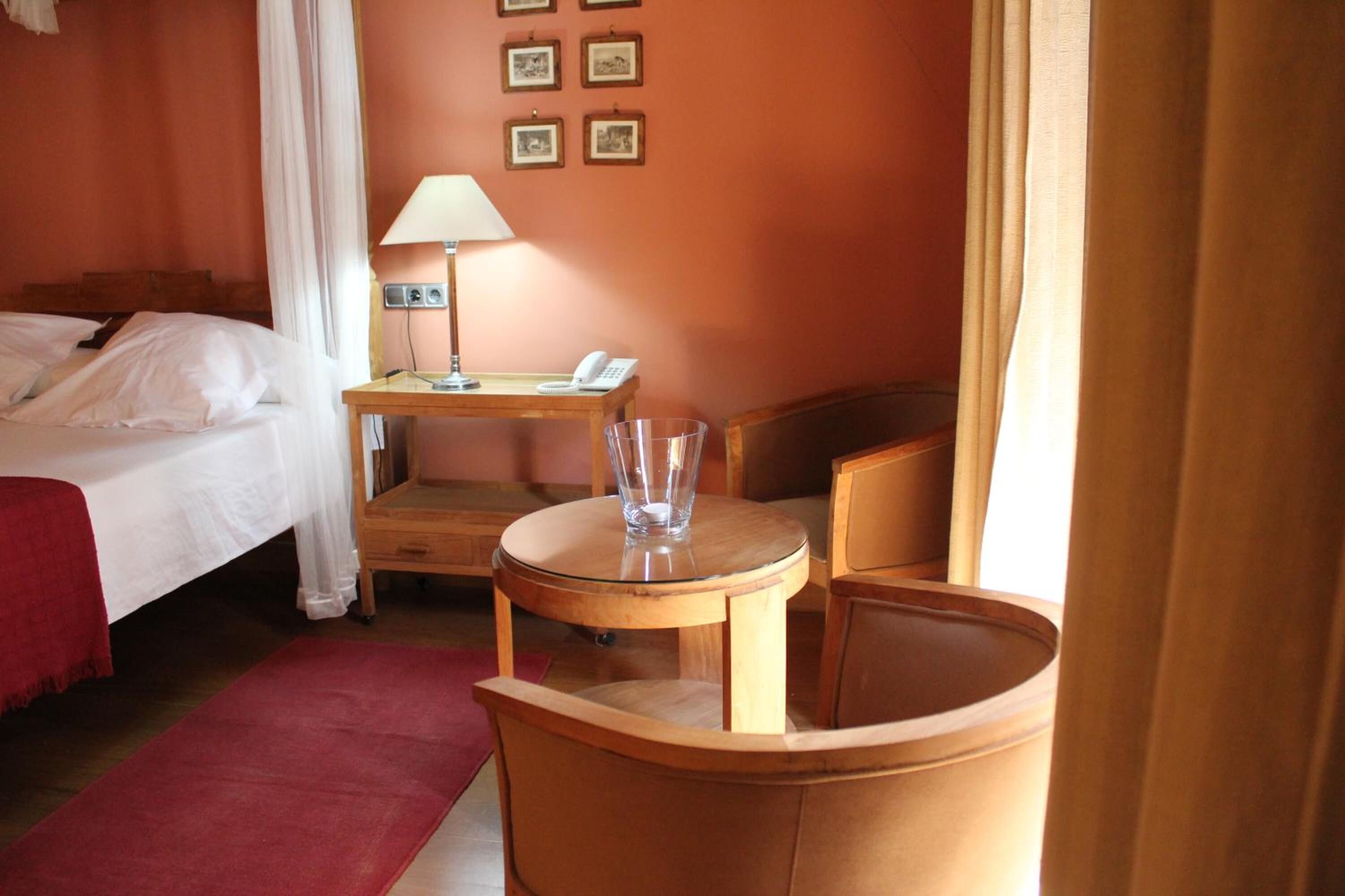 A hotel room with a bed, a desk with a lamp, a chair, and a small table with a glass.