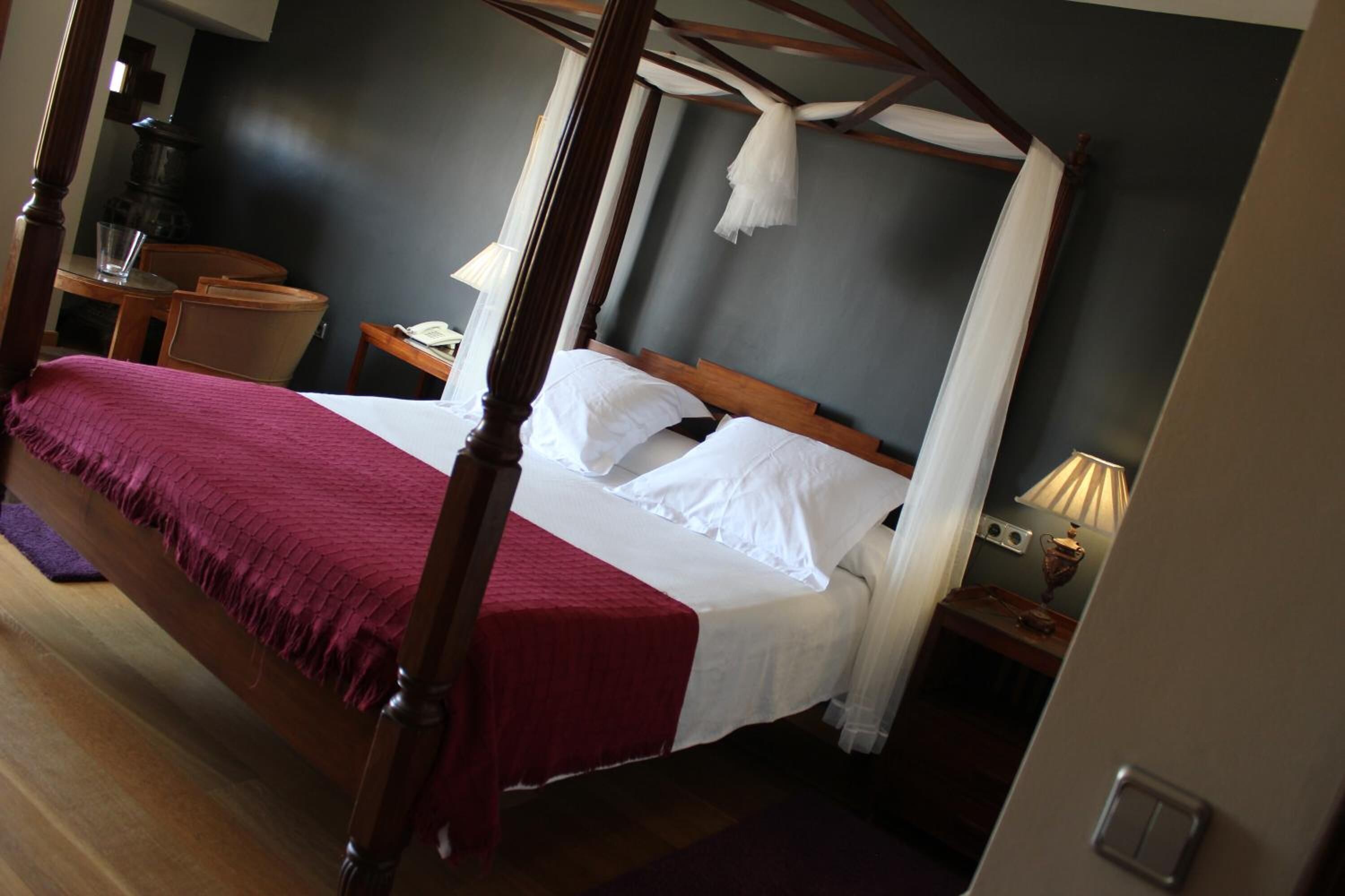 A four-poster bed with white linens, a wooden headboard, and a red throw blanket. There is a bedside table with a lamp and a telephone. The room has a dark wall and a wooden floor.