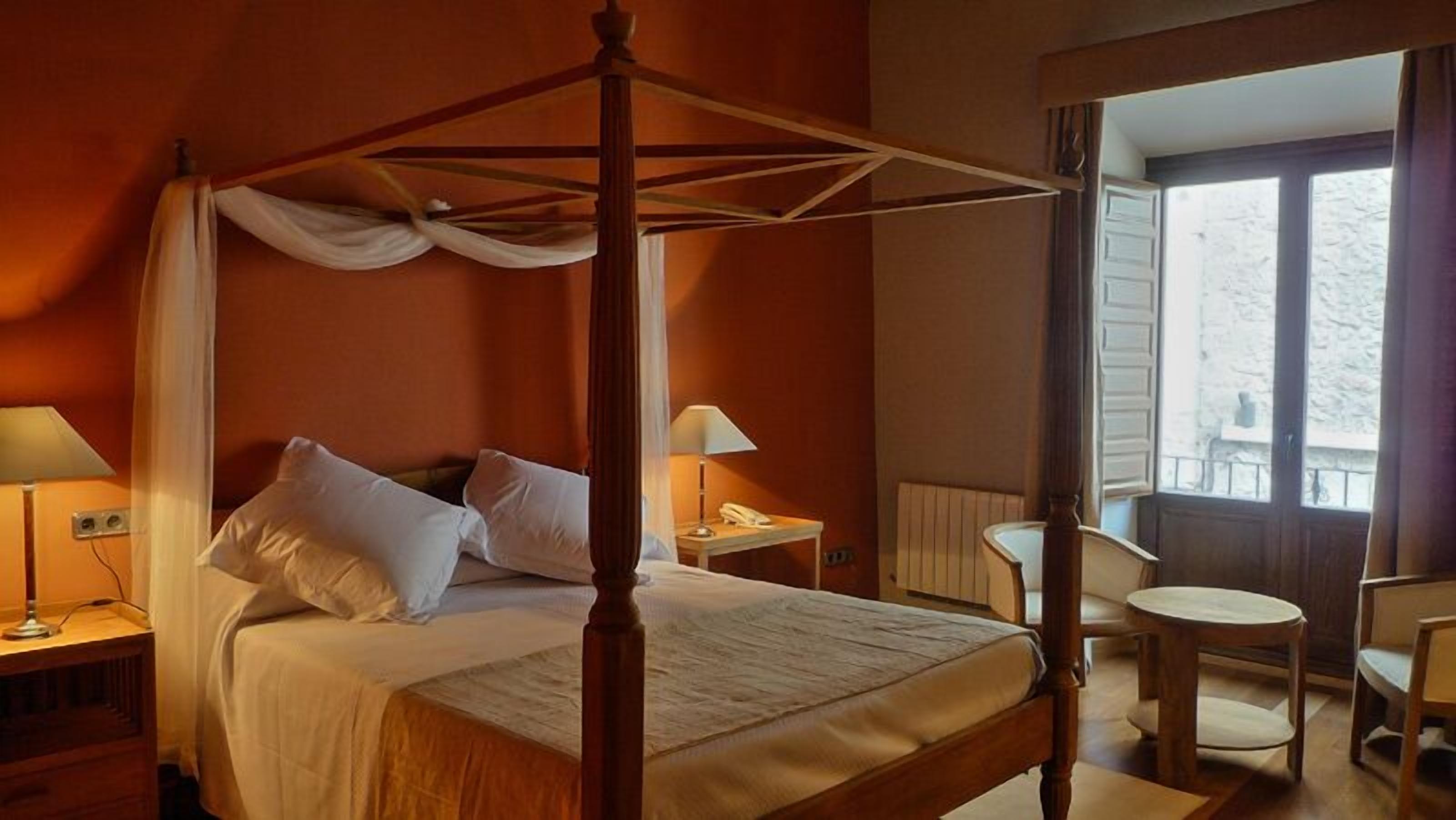 A four-poster bed with white linens, a bedside table with a lamp, a small round table with a chair, and a view of the outside through a window.