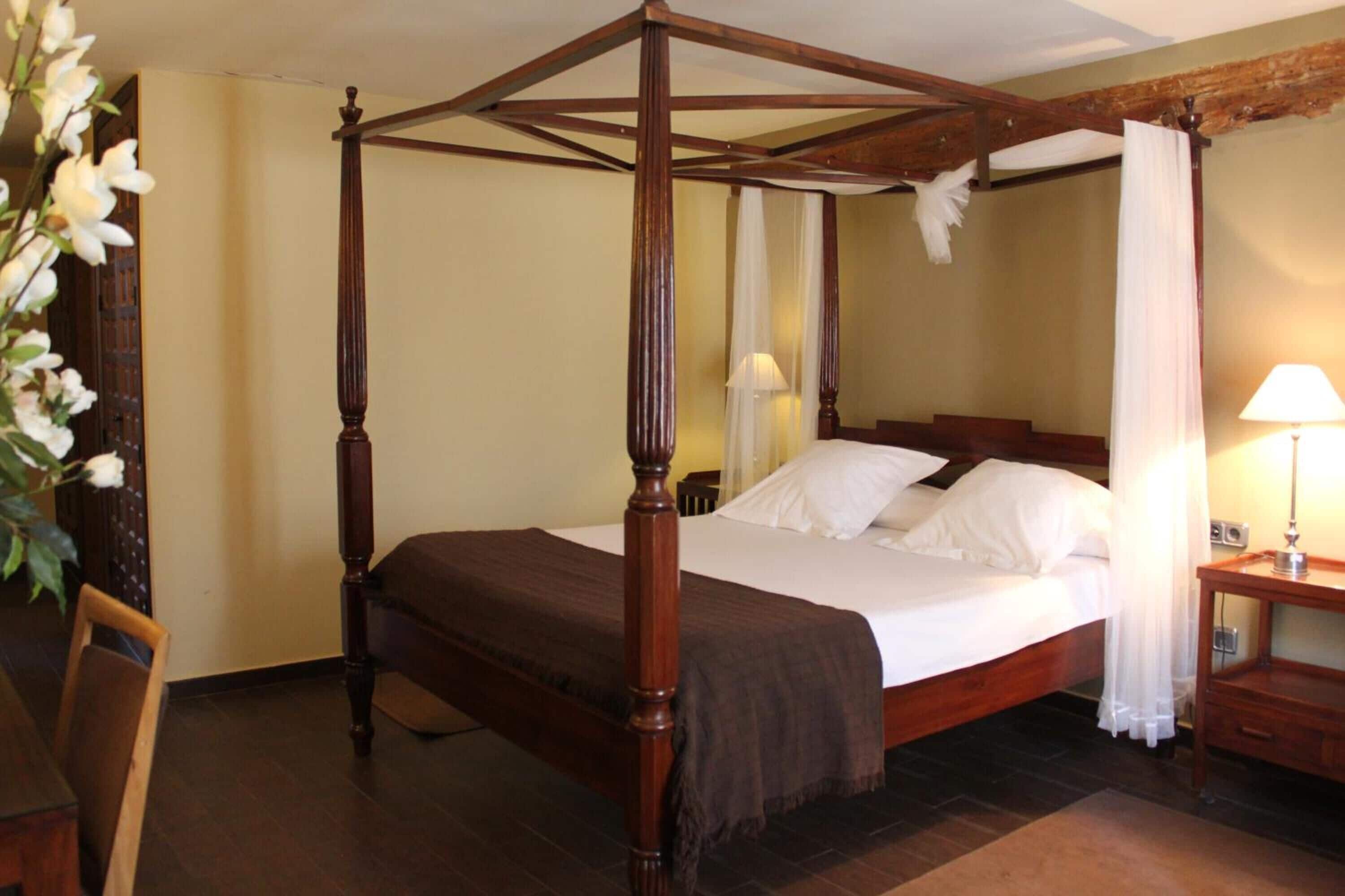 Una cama con dosel de madera, ropa de cama blanca y una mesita de noche con lámpara.