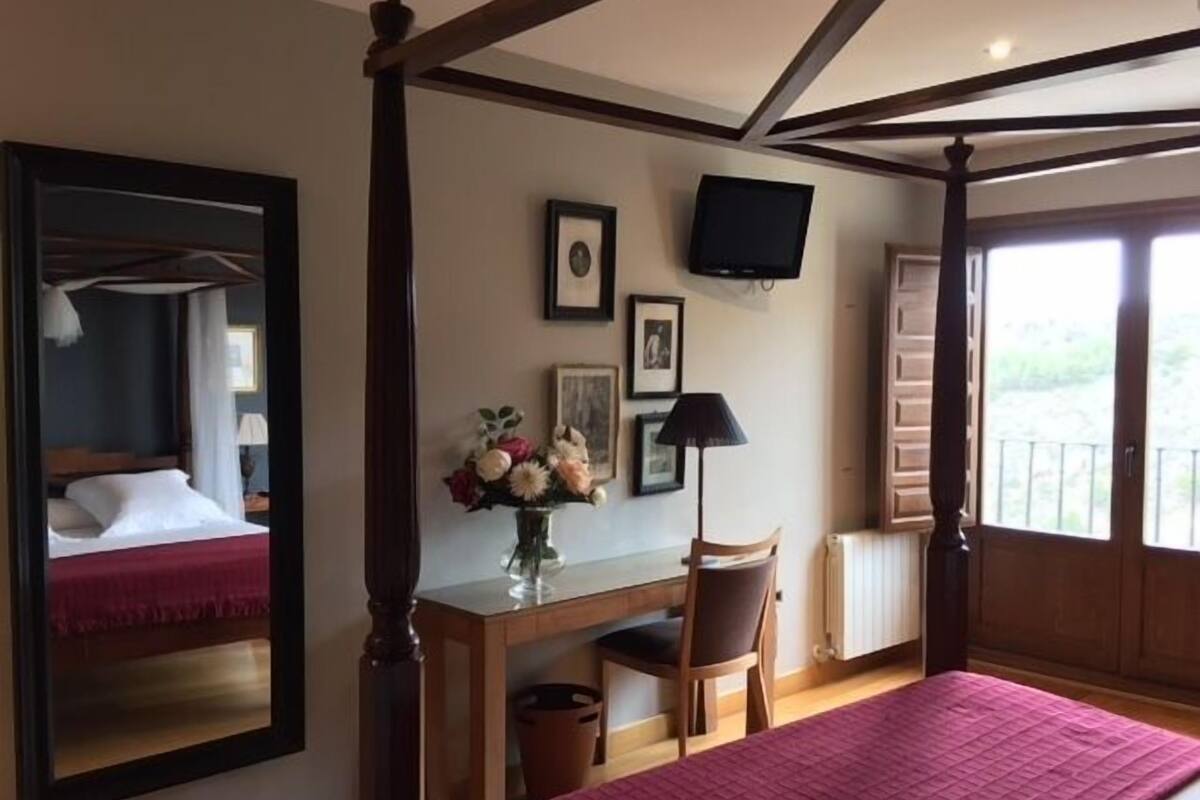 Un dormitorio con cama con dosel, un escritorio con silla, un televisor en la pared y una ventana con vistas a zonas verdes.
