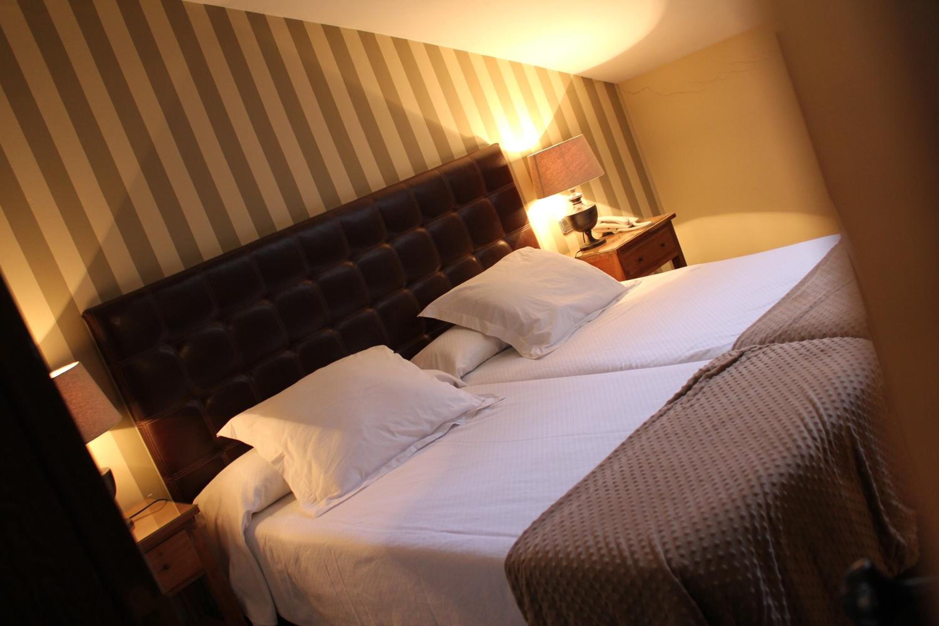 Een hotelkamer met een groot bed, twee kussens, een nachtkastje met een lamp en gestreept behang.