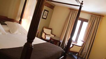 A four-poster bed with a wooden frame, a small table, and a chair in a room with beige walls and a window with curtains.