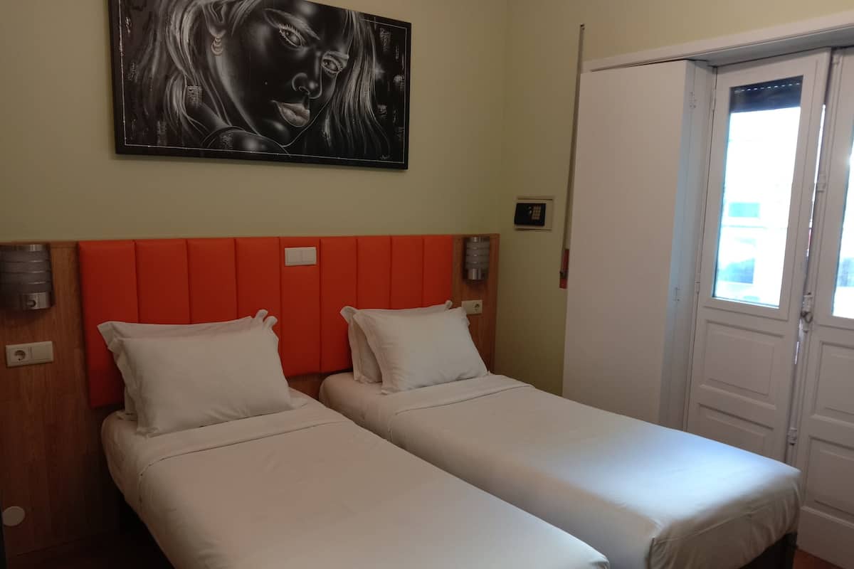 A hotel room with two single beds, a red headboard, a painting on the wall, and a door with a window.