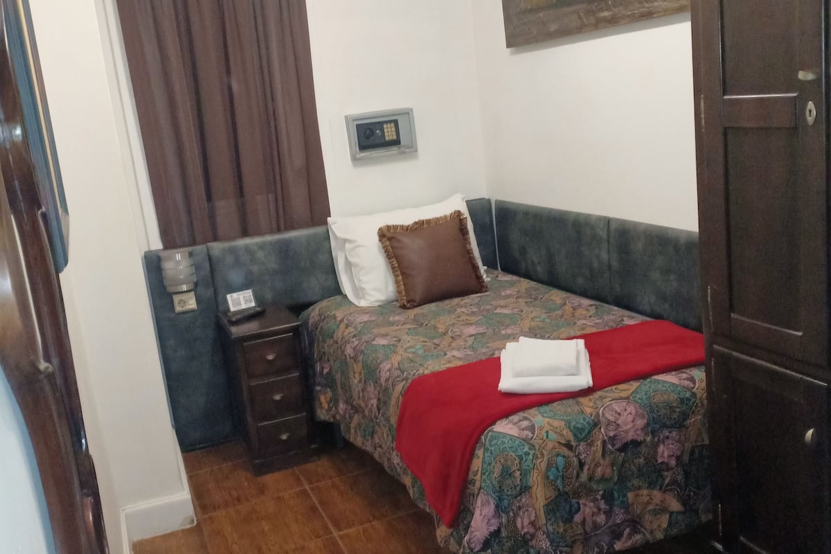 A bedroom with a bed, a nightstand, a wardrobe, a painting, and an air conditioning unit.