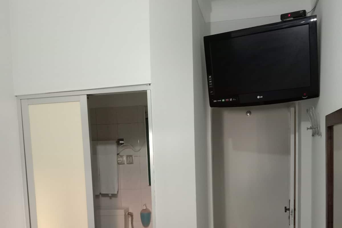 A bathroom with a toilet, sink, and shower. There is a TV mounted on the wall and a small stool with cups.