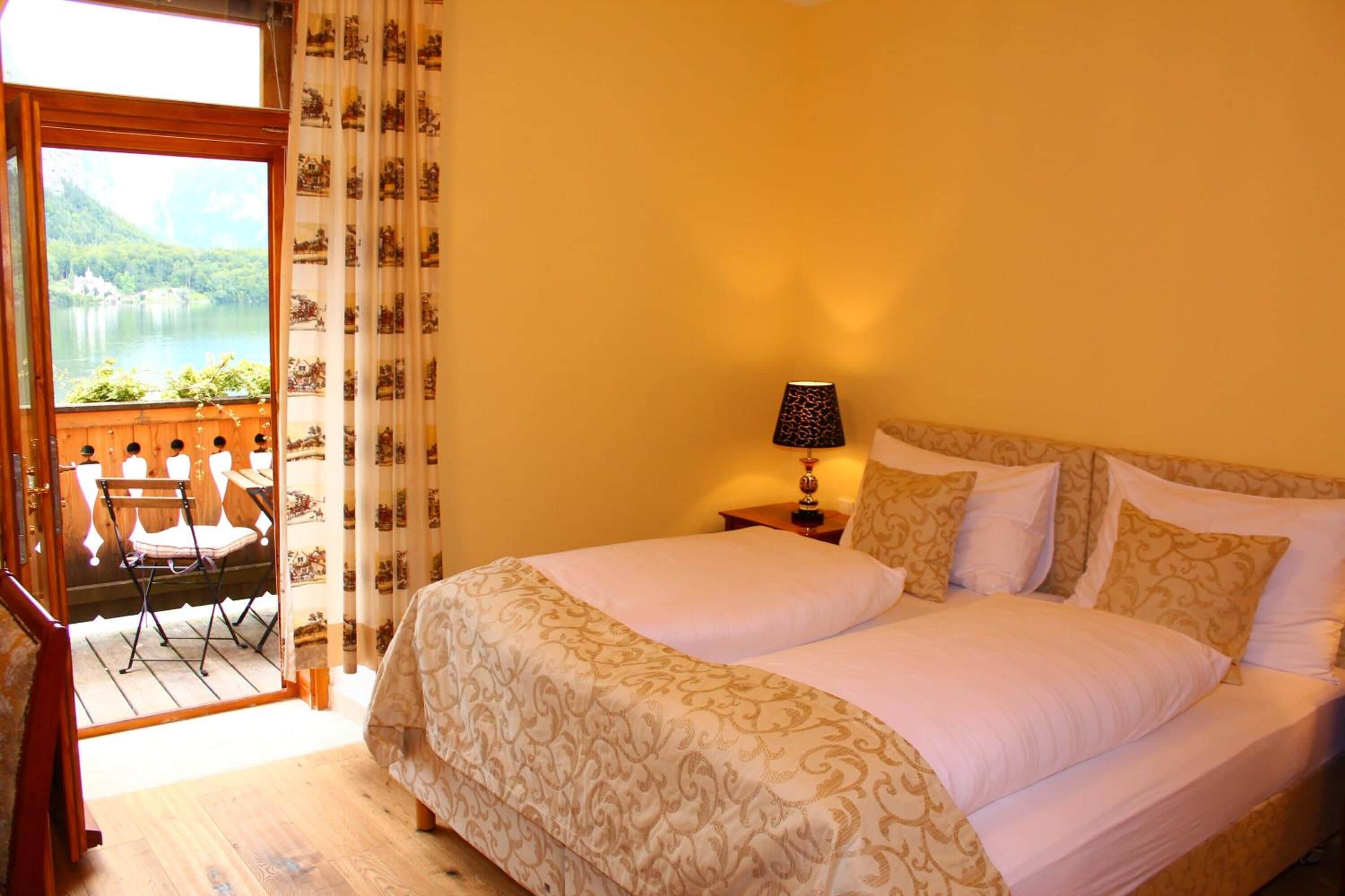 A hotel room with a large bed, a nightstand with a lamp, a view of a lake through a window, and a balcony with chairs.