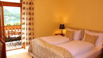 A hotel room with a large bed, a nightstand with a lamp, a view of a lake through a window, and a balcony with chairs.