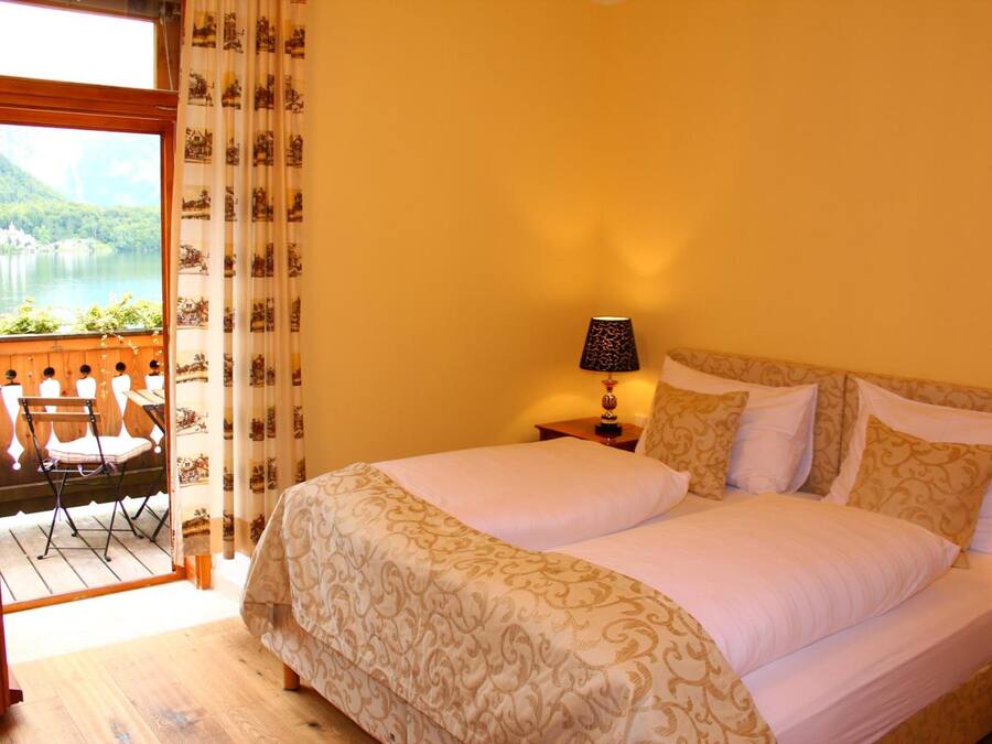 A hotel room with a large bed, a nightstand with a lamp, a view of a lake through a window, and a balcony with chairs.