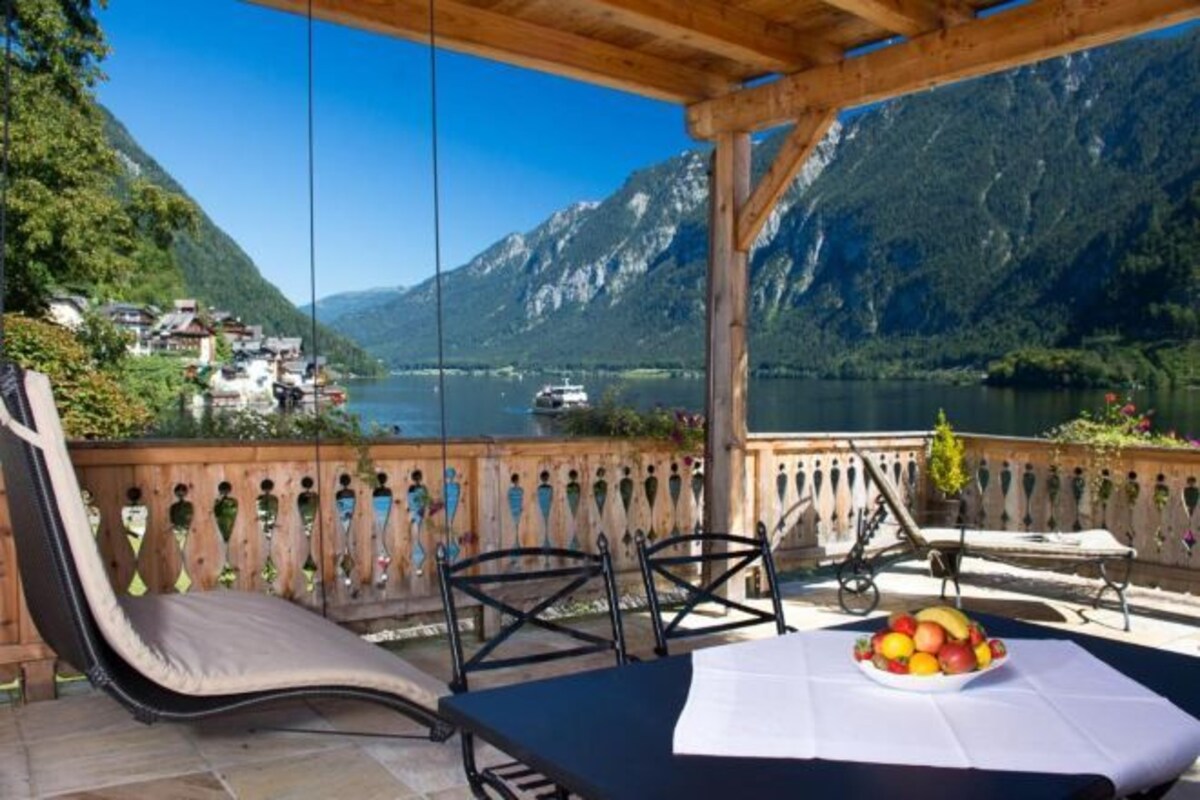 A terrace with a table set for dining, a chair, and a view of a lake with mountains in the background.