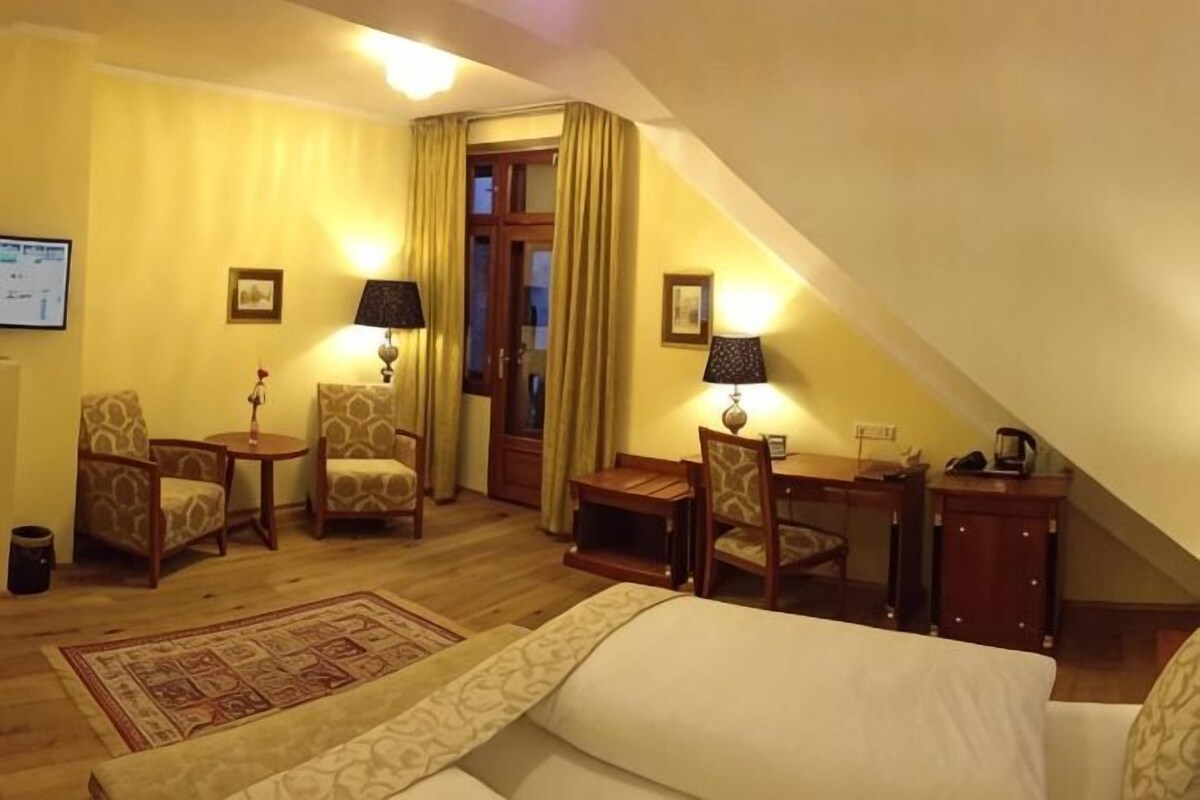 A hotel room with a bed, two chairs, a small table, a desk, and a lamp.