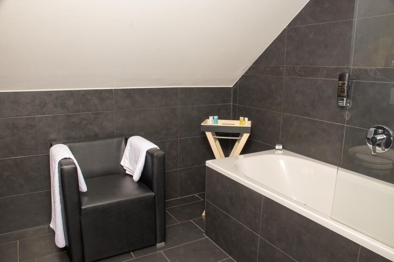 A modern bathroom with a black bathtub, a black cushioned chair, and a wooden shelf with toiletries.