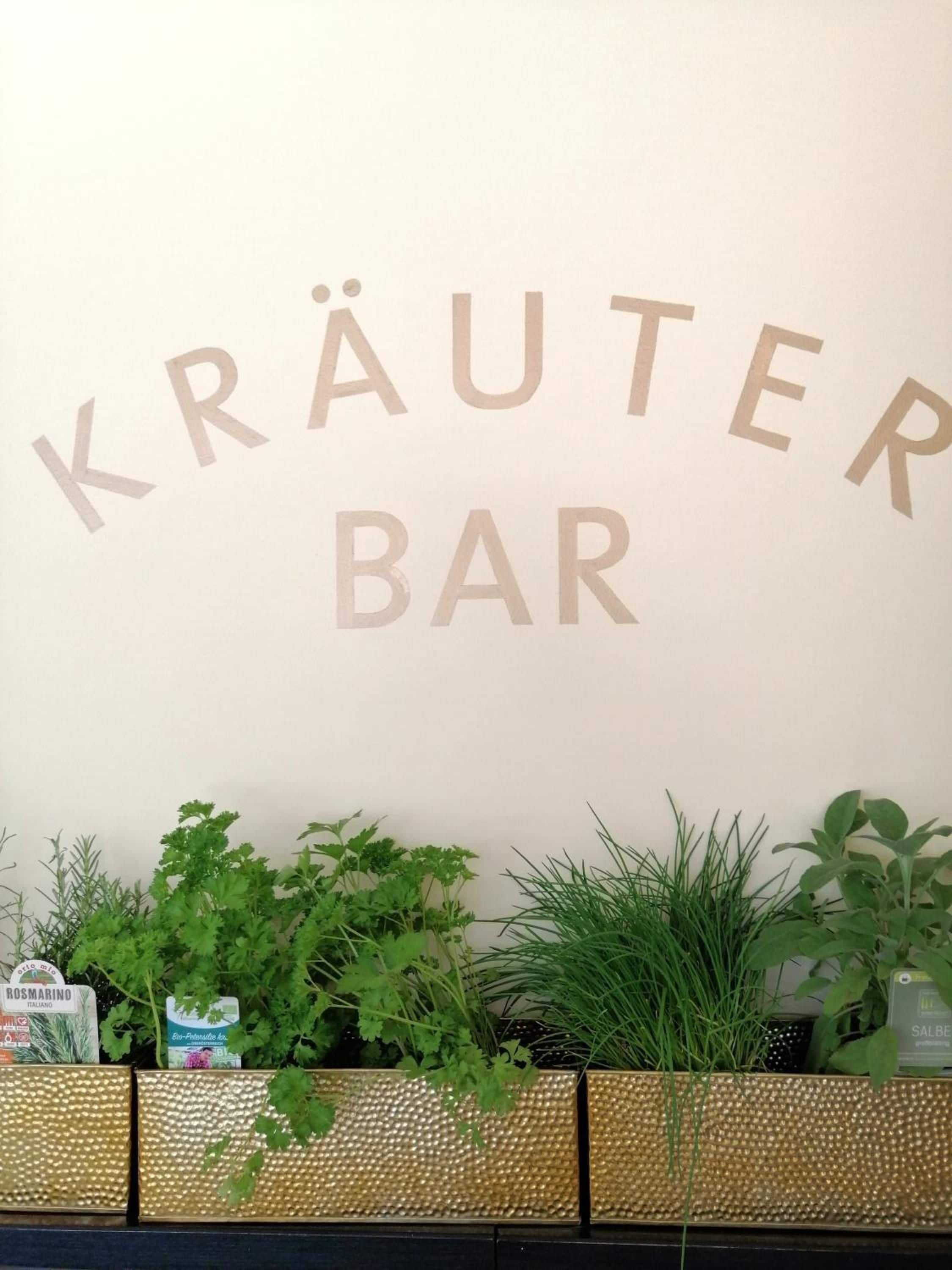 A wall with the text 'KRÄUTER BAR' and a planter with various green plants.