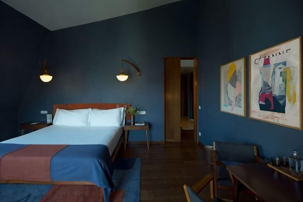 A bedroom with a bed, bedside tables, a desk, and chairs. There are framed artworks on the wall.