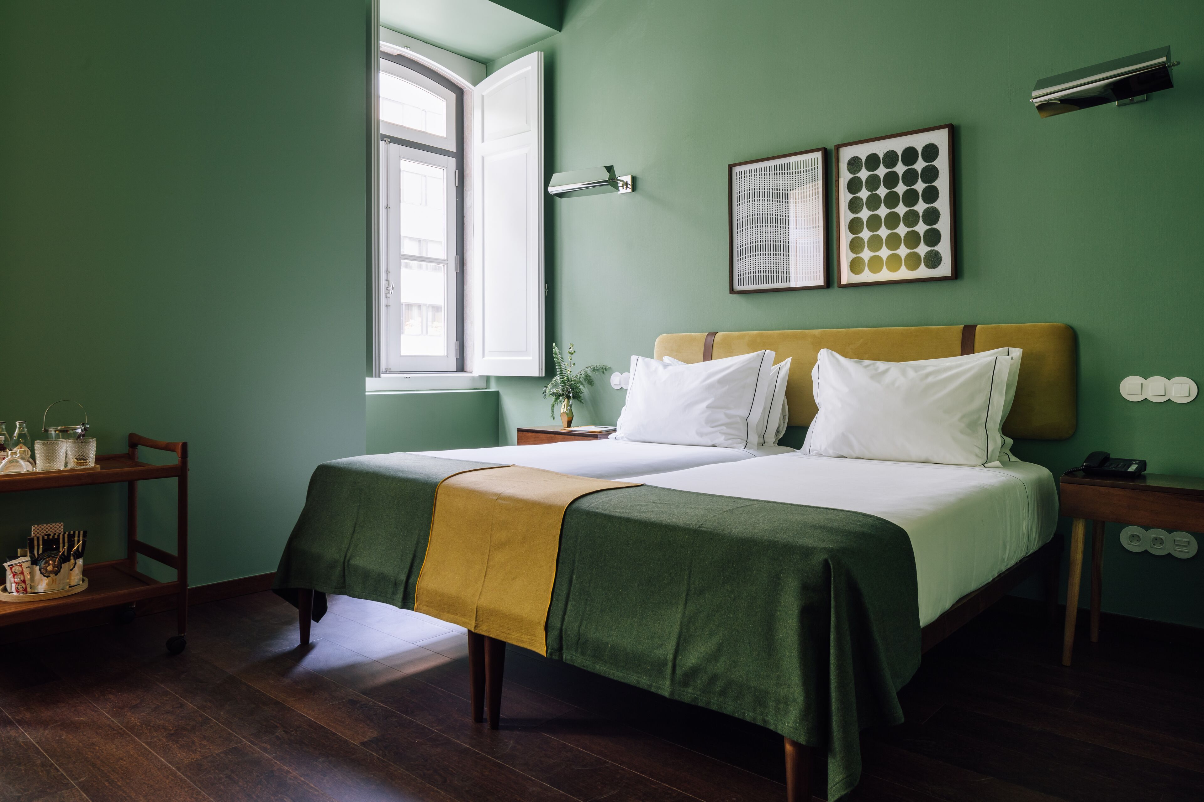 Uma cama de casal com colcha verde e amarela, uma mesinha de cabeceira com abajur e uma janela com vista para o exterior.