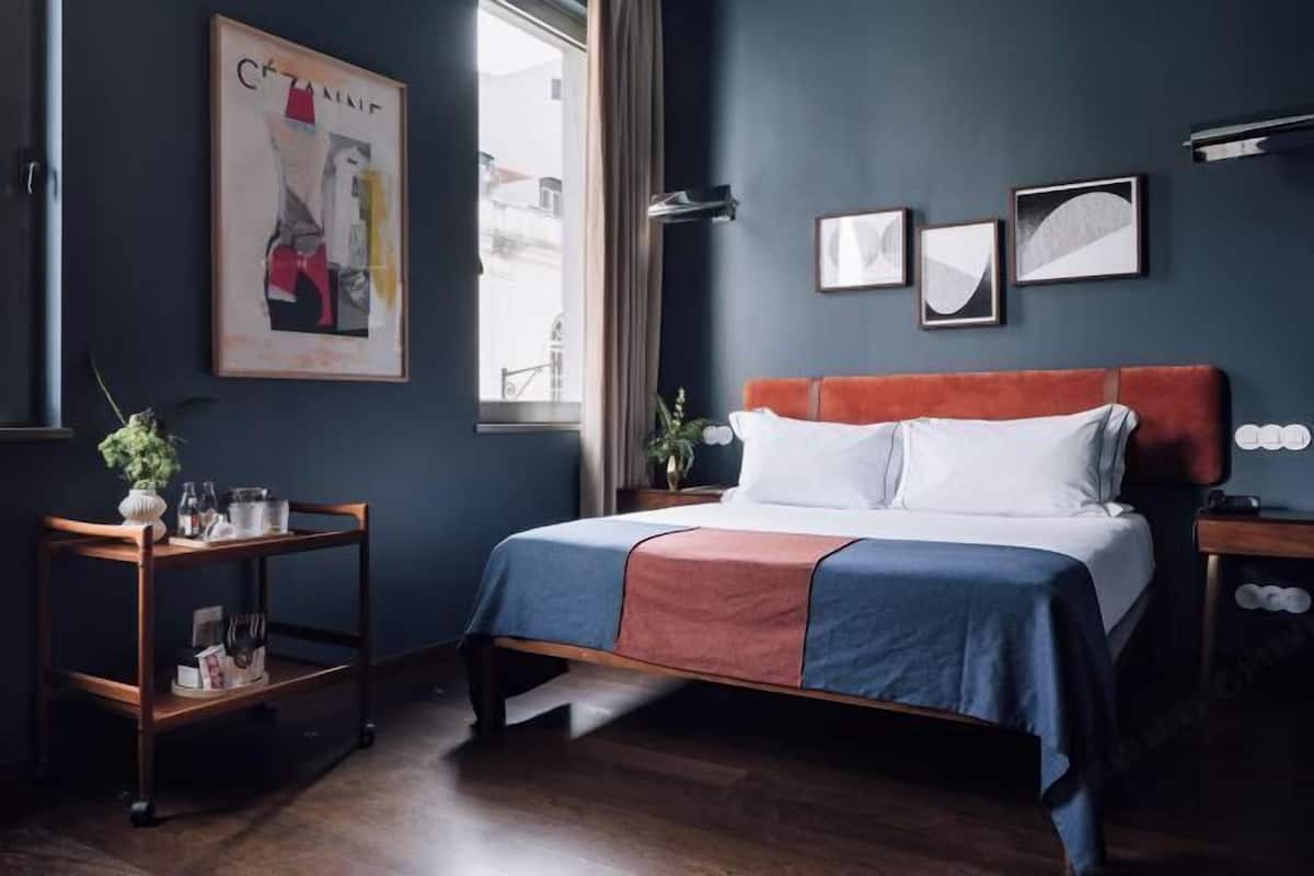 A bedroom with a bed, a nightstand, a wooden side table, a window, and wall art.