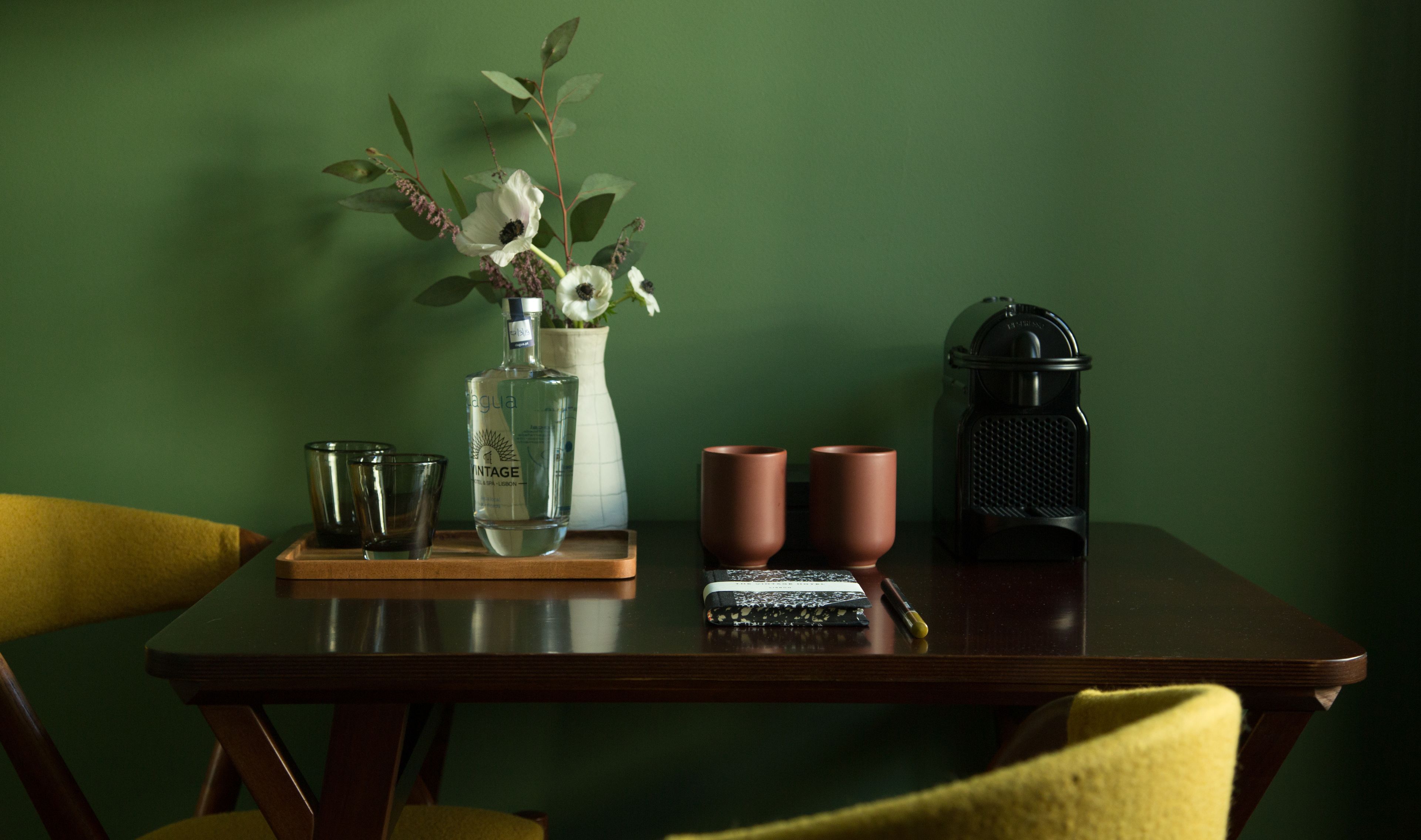 A wooden table with a clear glass bottle, two brown cups, a small black object, and a vase with flowers against a green wall.
