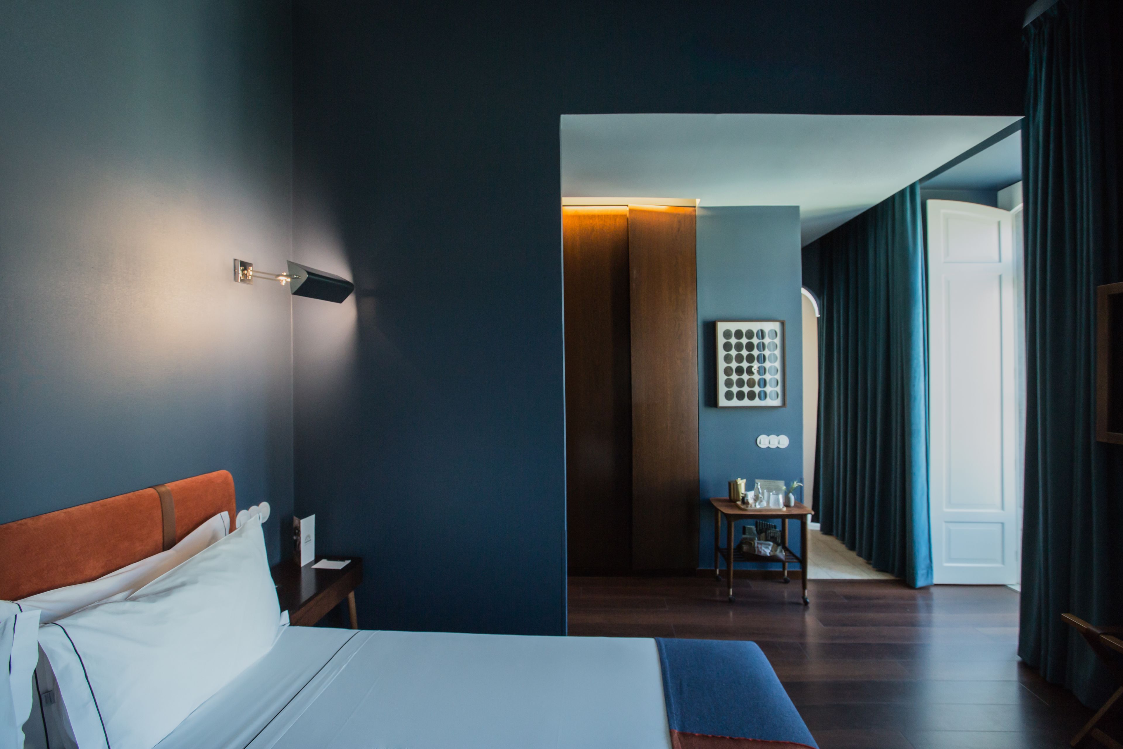 Quarto de hotel moderno com cama bem arrumada, mesa de cabeceira, luminária na parede e vista para o quarto vizinho.