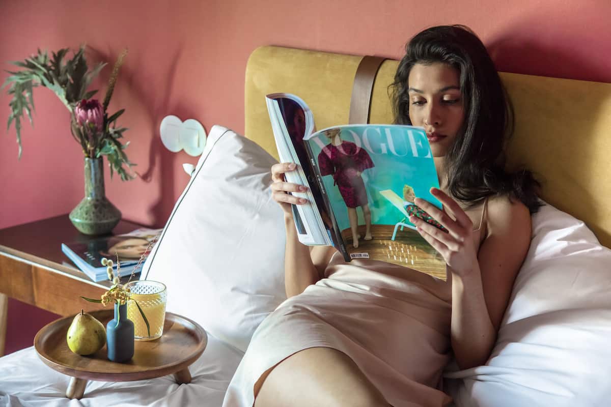 A person lying on a bed reading a magazine, with a small table beside the bed holding a vase with flowers and a glass of drink.