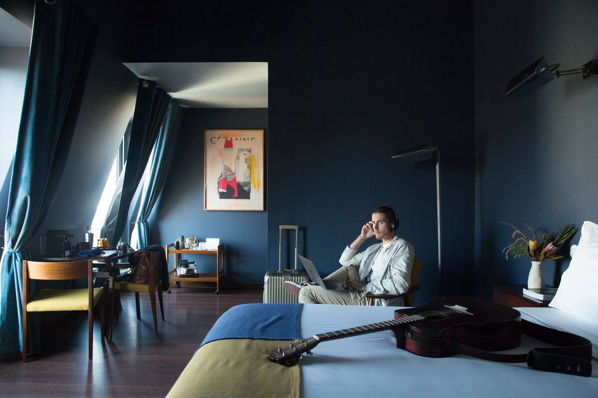 A hotel room with a bed, a guitar, a laptop, a desk with a chair, and a window with curtains.