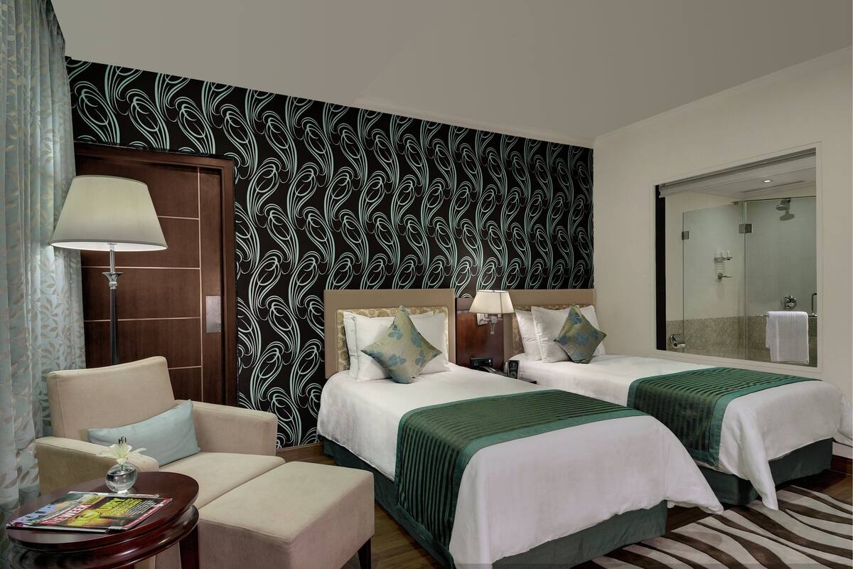 Quarto de hotel com duas camas, um sofá, uma mesa pequena com uma revista e papel de parede estampado.