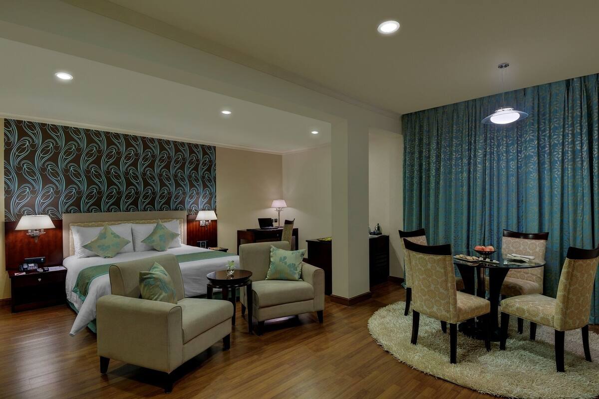Quarto de hotel com cama, duas poltronas, uma mesinha, escrivaninha e um espaço de jantar com cadeiras.