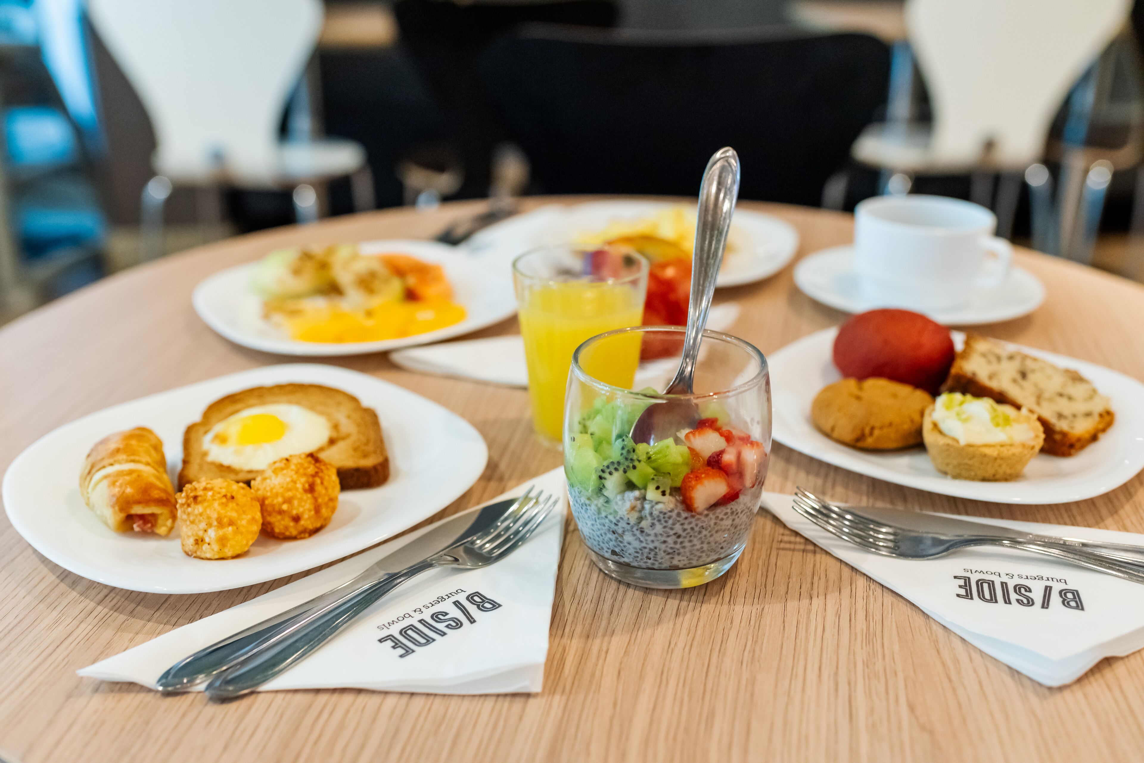 A breakfast setting with plates of eggs, toast, and fruit, along with a glass of orange juice and a chia seed pudding.