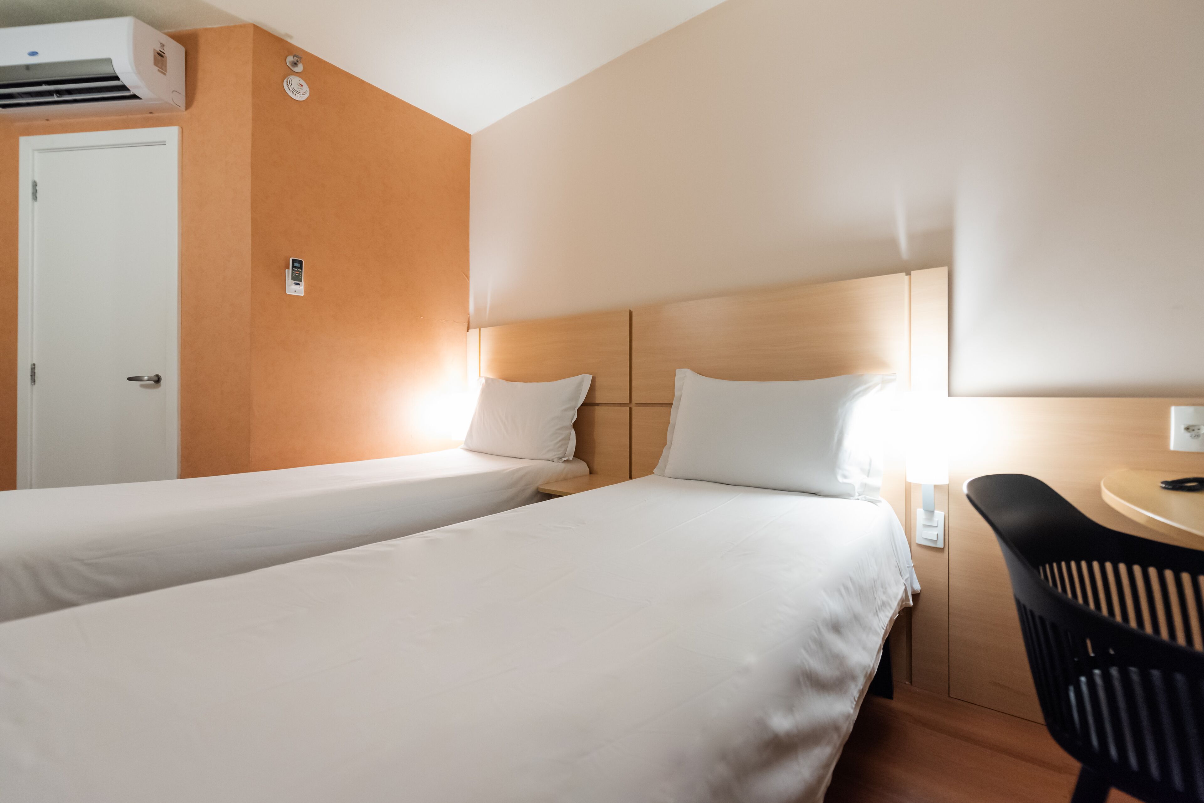 A hotel room with two single beds, a desk with a chair, an air conditioning unit, and a door.