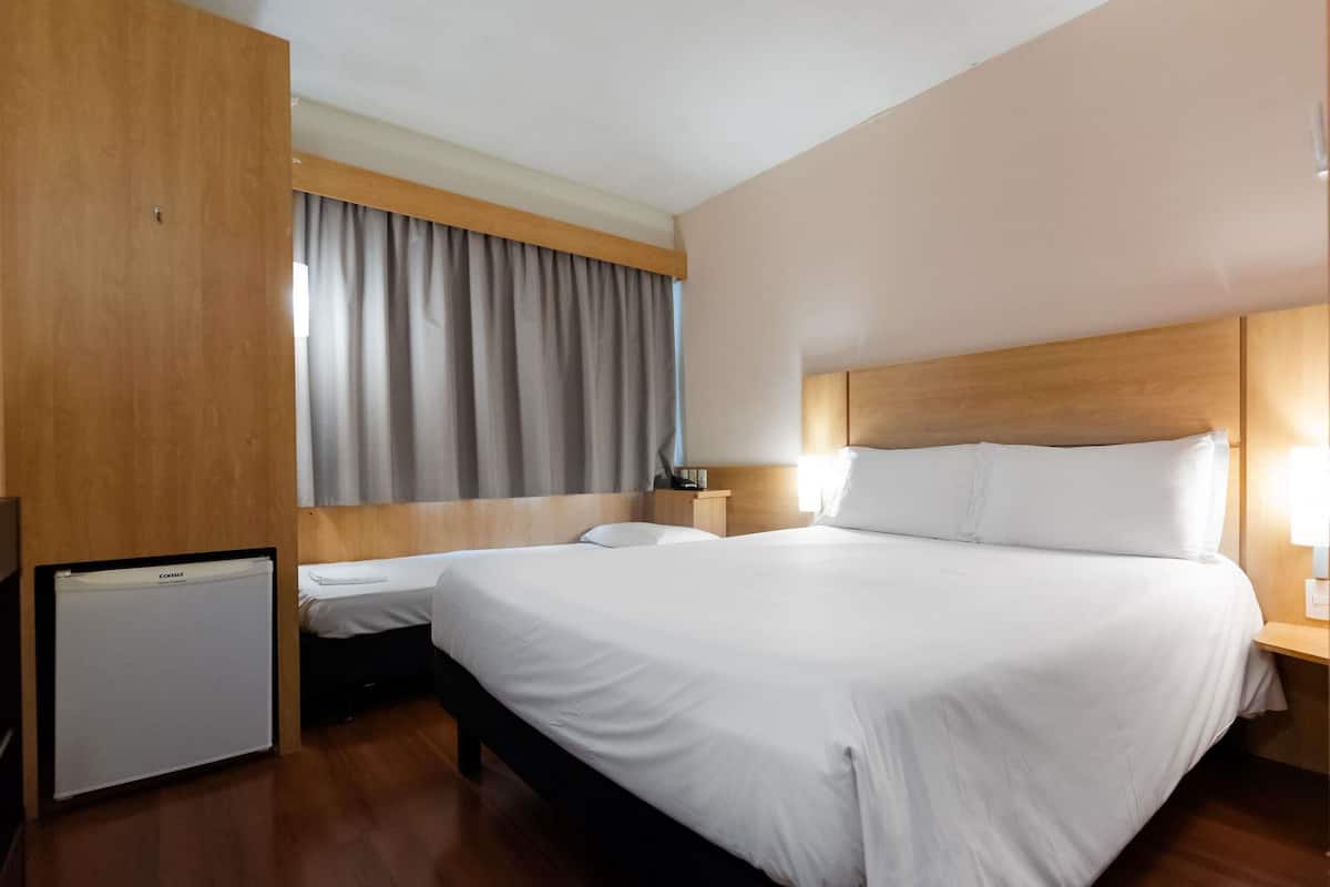 A hotel room with a large bed, a bedside table, a lamp, a small desk with a computer, and a window with curtains.