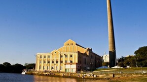 A large industrial building with a tall chimney, a water body in the foreground, and a grassy area with a small hill. - ibis Porto Alegre Moinhos de Vento (Porto Alegre)