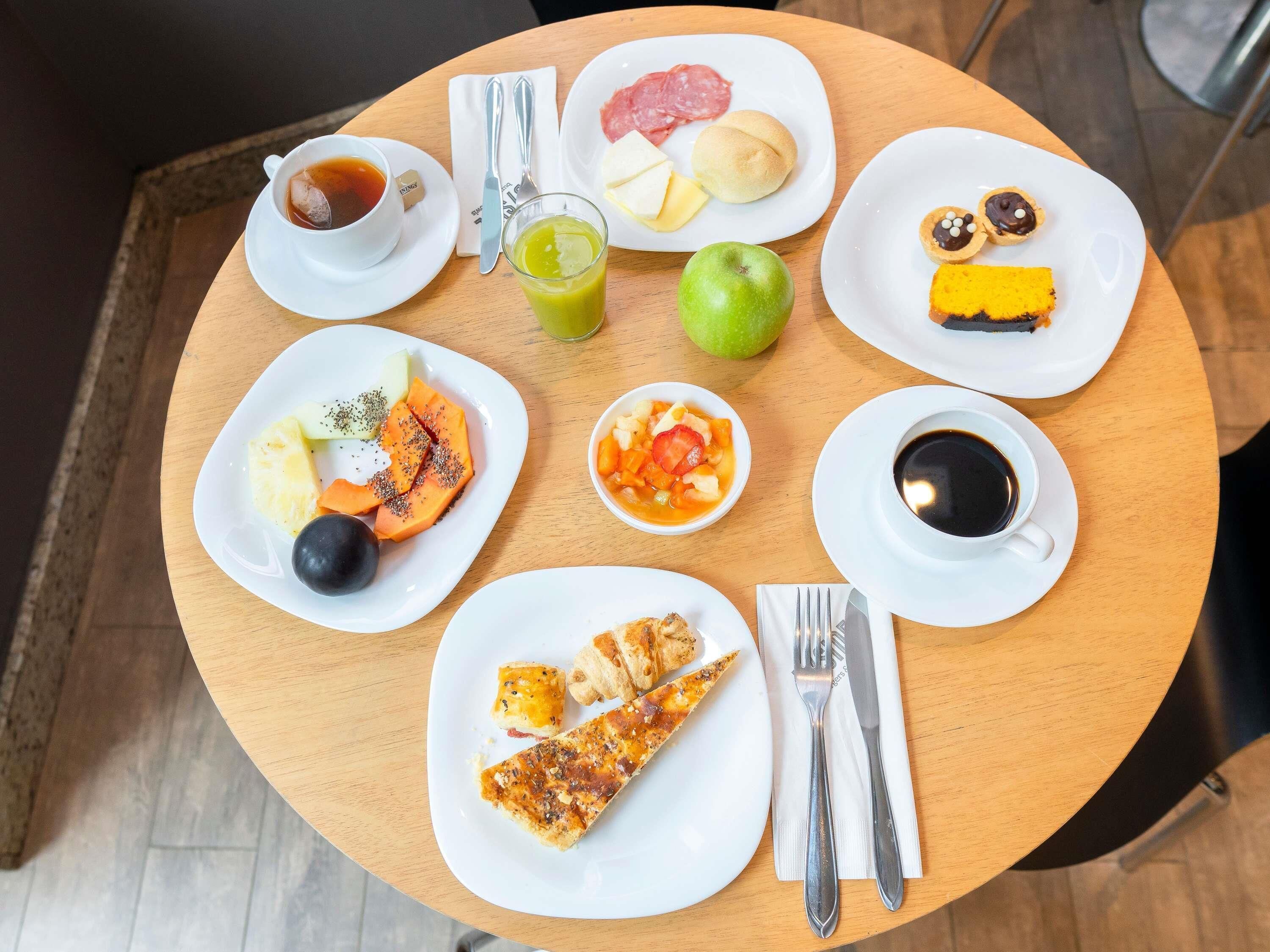Pyöreä puinen pöytä, jonka päällä on erilaisia aamiaistarvikkeita, kuten vihreä omena, pizzapala sekä lautanen, jossa on monenlaisia hedelmiä ja kuppi kahvia.