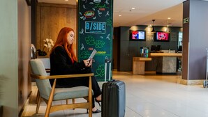 A woman sitting on a chair with a suitcase beside her, in a modern lobby with a 'B/SIDE burgers & bowls' sign and a reception area in the background. - ibis Porto Alegre Moinhos de Vento (Porto Alegre)