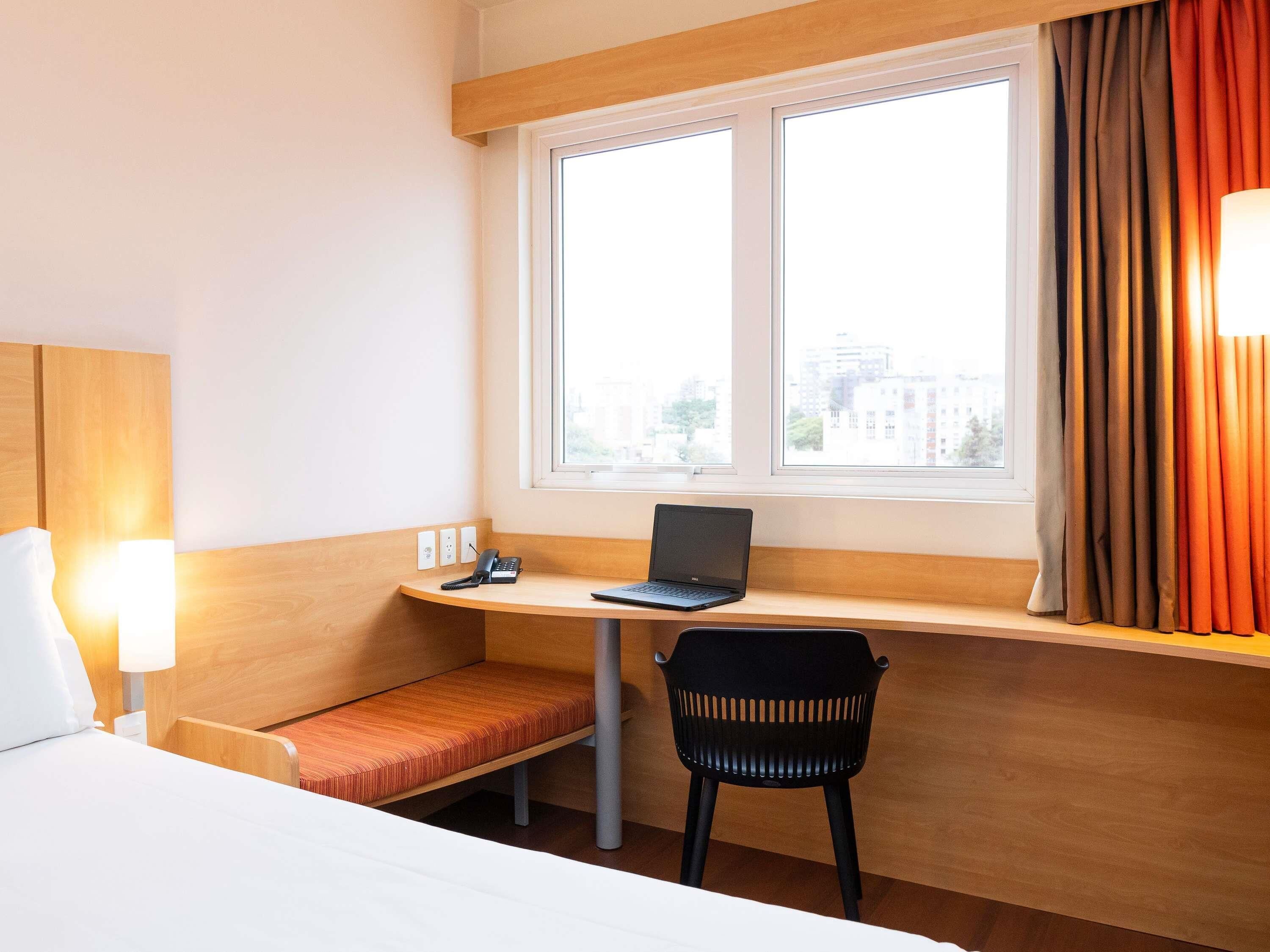 A hotel room with a bed, a desk with a laptop, a chair, and a window with curtains.
