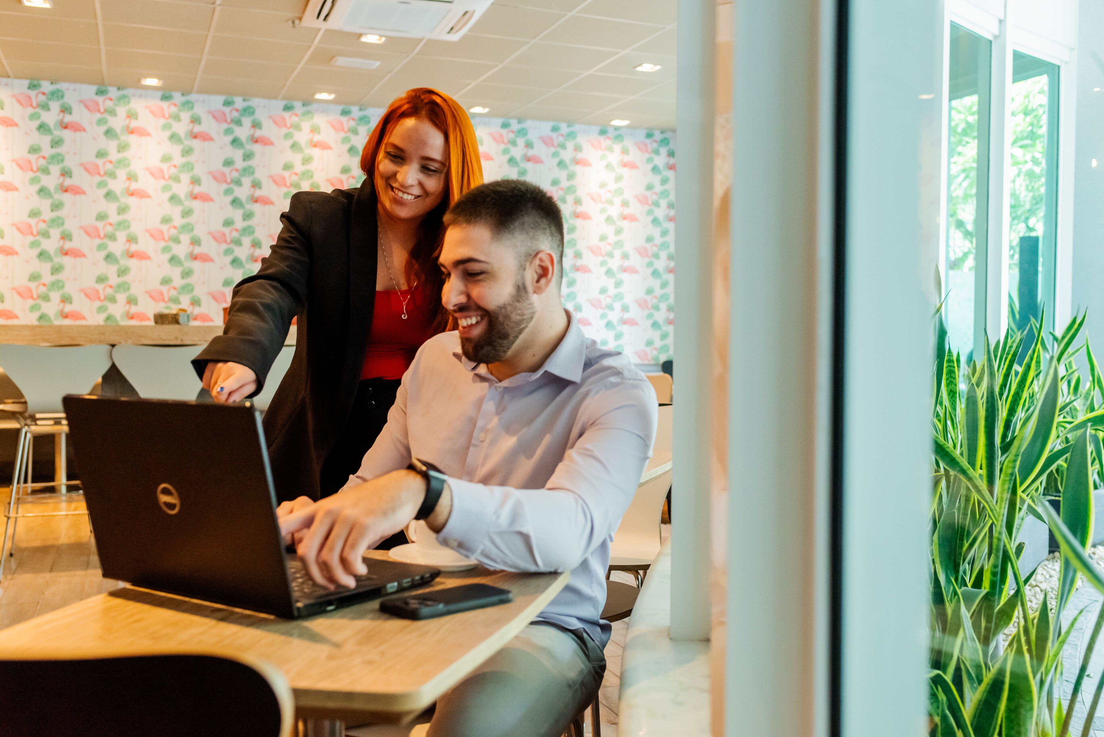 Un homme et une femme sont assis à une table devant un ordinateur portable, dans une pièce dont les murs sont recouverts d’un papier peint à motifs floraux et où se trouve une plante dans un coin.
