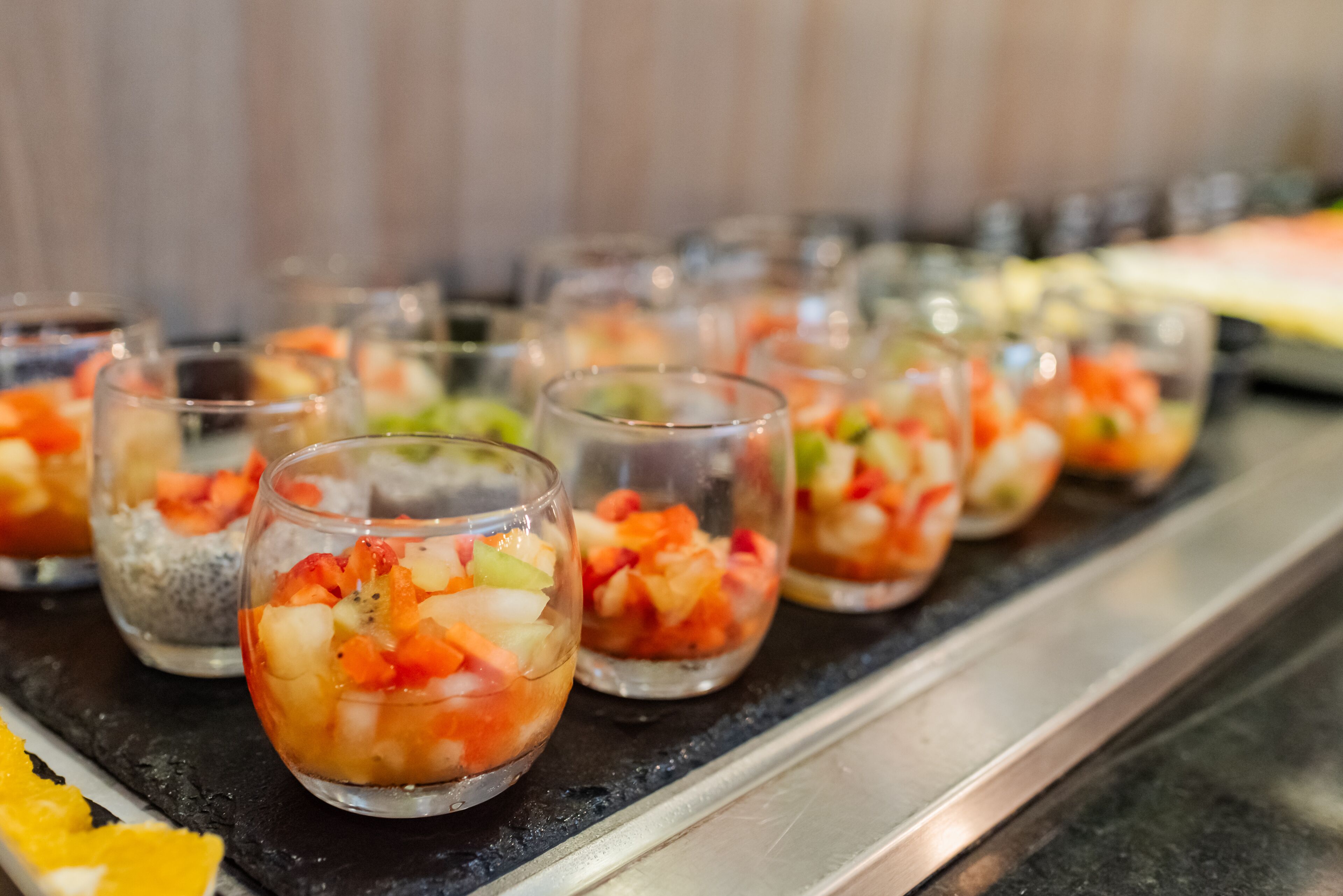 Una hilera de vasos pequeños con una mezcla de ensalada de frutas, sobre una encimera negra.