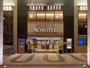 Entrada de un hotel Novotel con un mostrador de recepción, plantas en macetas y una zona de estacionamiento para bicicletas.