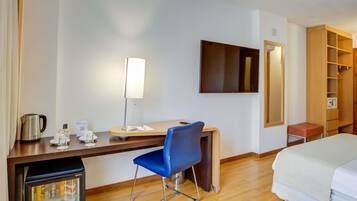 A hotel room with a bed, a desk with a chair, a refrigerator, a coffee maker, a mirror, and a television.