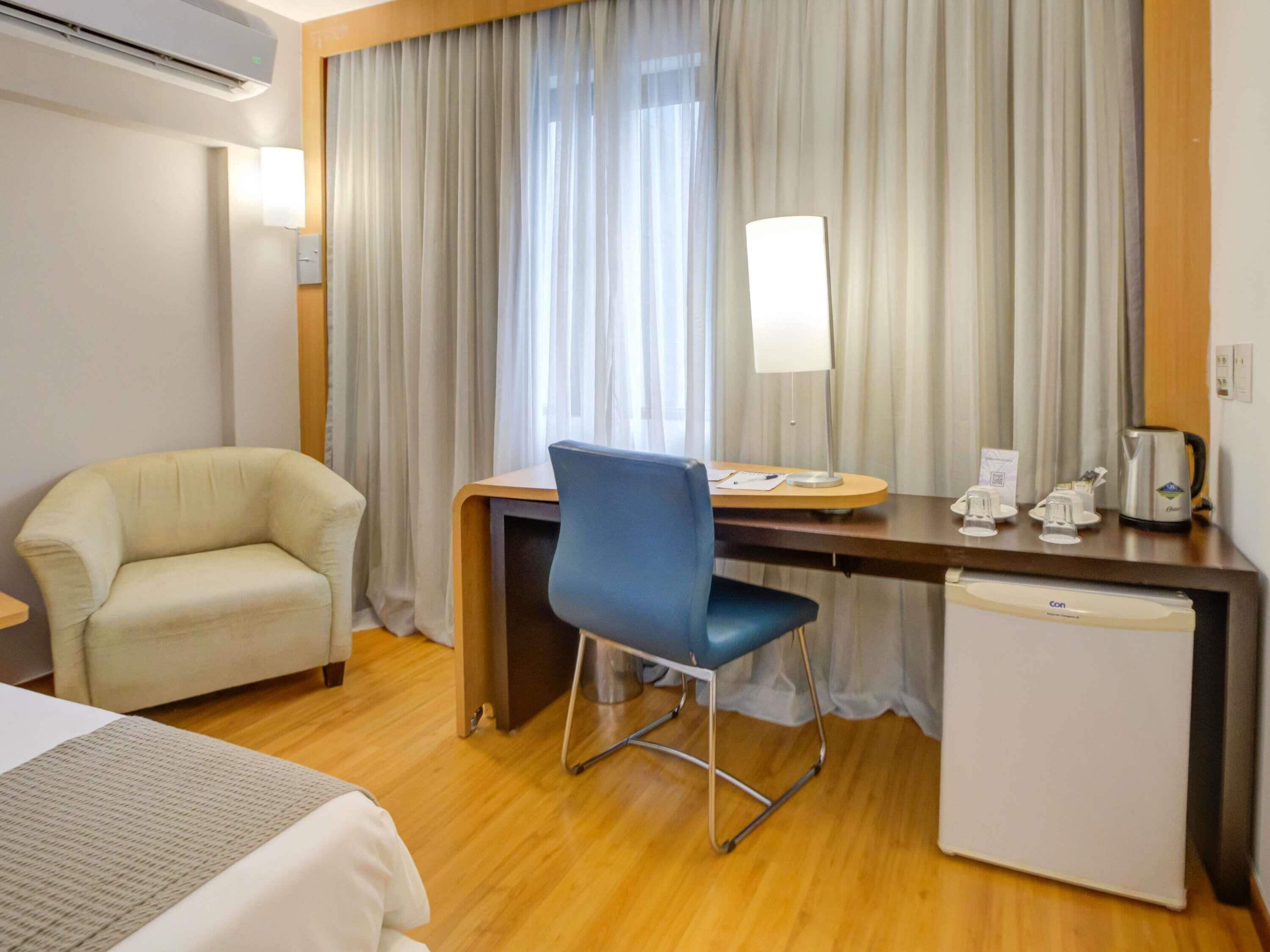 A hotel room with a bed, a desk with a chair, a coffee maker, and a refrigerator.