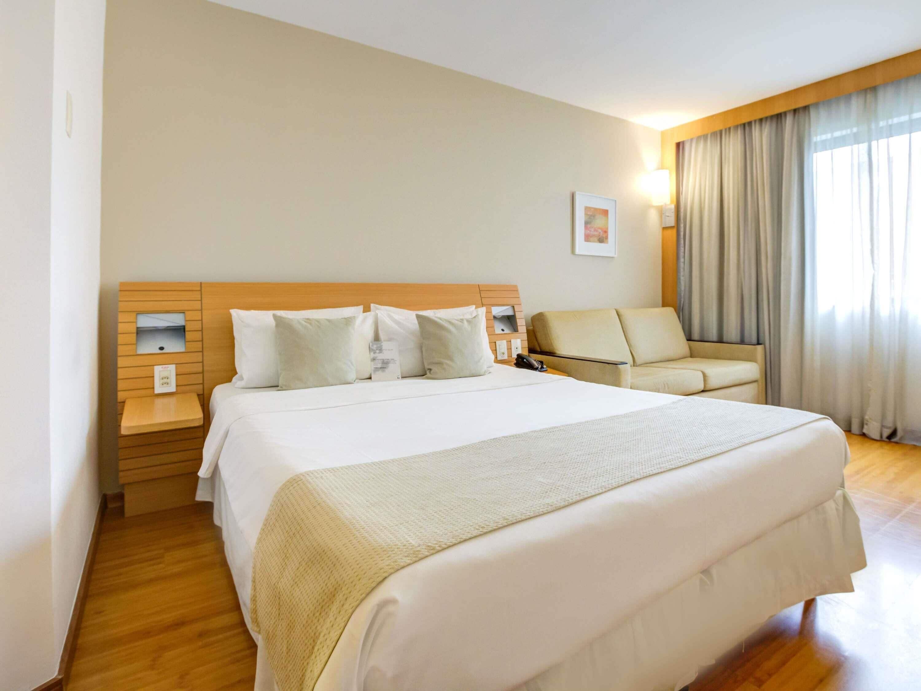 A hotel room with a large bed, a bedside table, a chair, and a window with curtains.