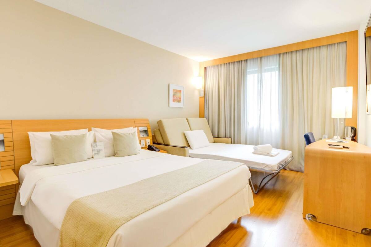 Habitación de hotel con dos camas, un escritorio y un ventanal con cortinas.
