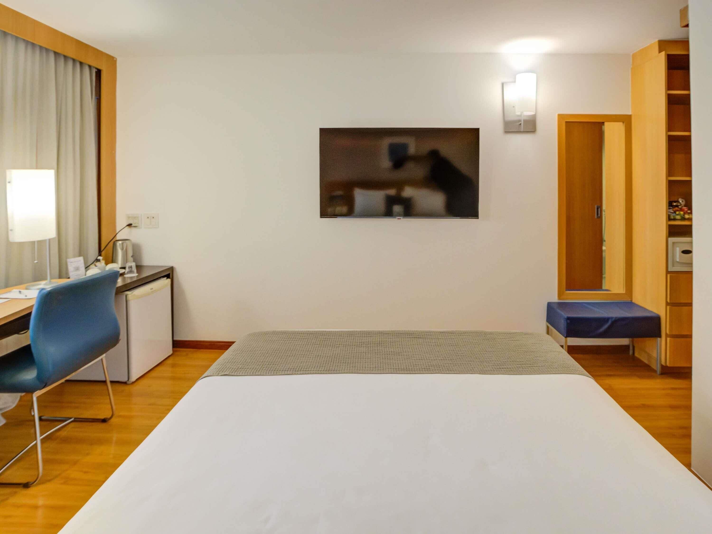 Habitación de hotel con cama, escritorio, silla, mesita de noche, lámpara y un cuadro en la pared.