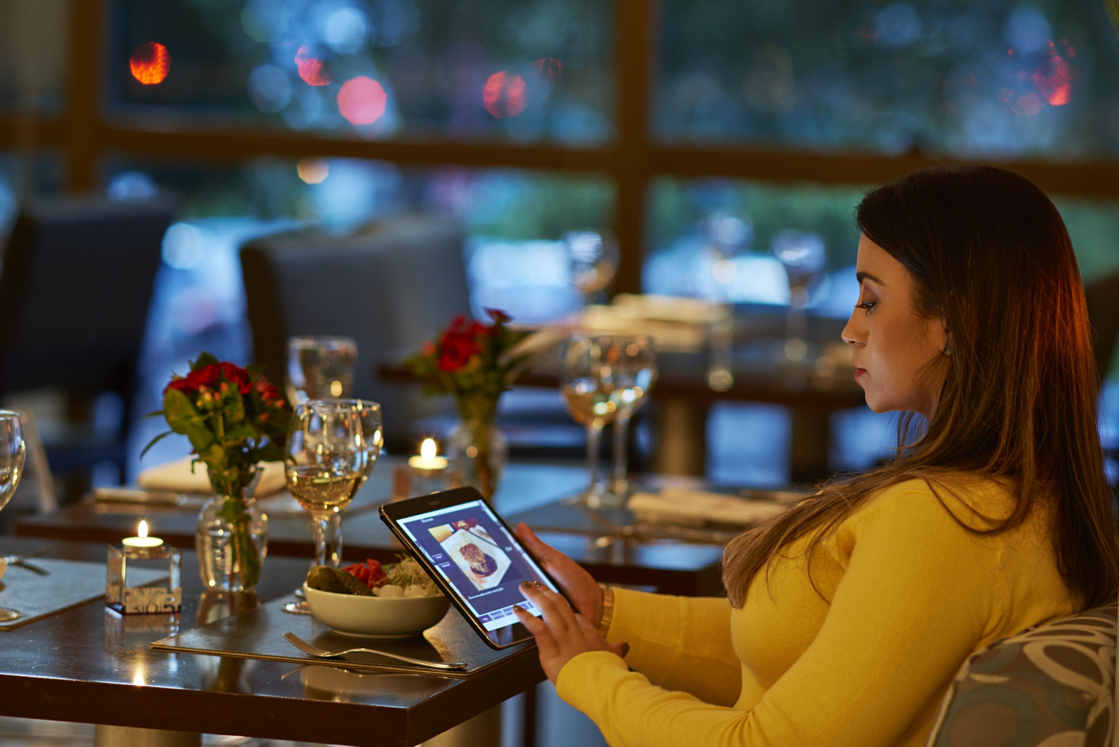 Una mujer sentada en una mesa de restaurante con una tableta, velas y un jarrón de flores.
