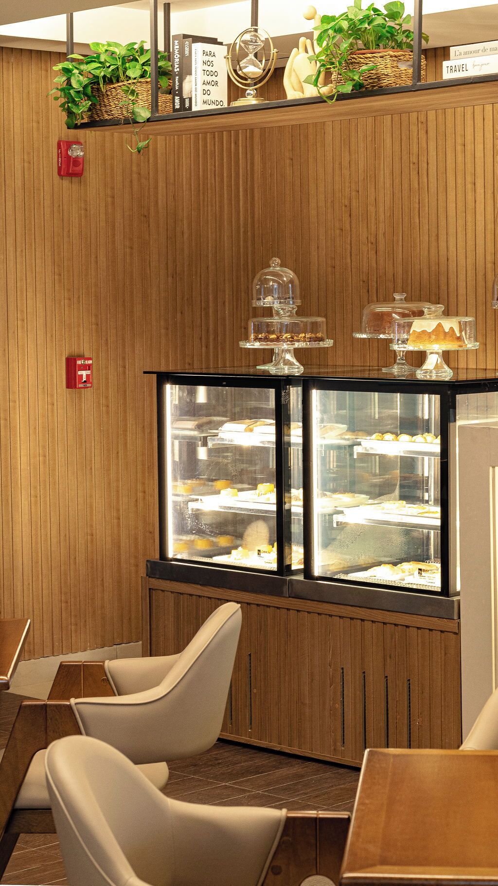 A restaurant with wooden paneling, a display case with desserts, and a shelf with plants and books.