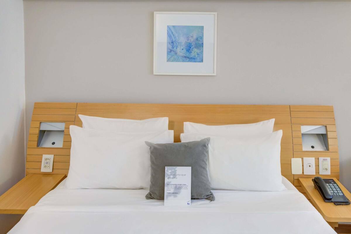 Habitación de hotel con cabecera de madera, ropa de cama blanca, almohada gris, un cuadro abstracto enmarcado, lámparas de noche y un teléfono en la mesita de noche.